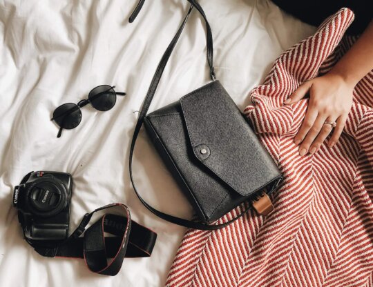 Black sunglasses, a black purse, and a DSLR camera lie on a bed comforter