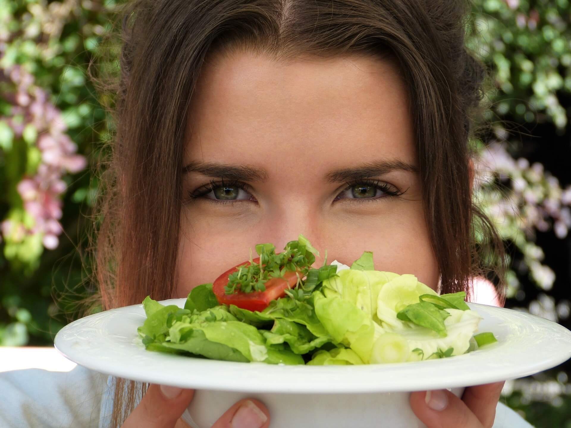 Young woman looking to camera holding a plate of salad in front of her face.